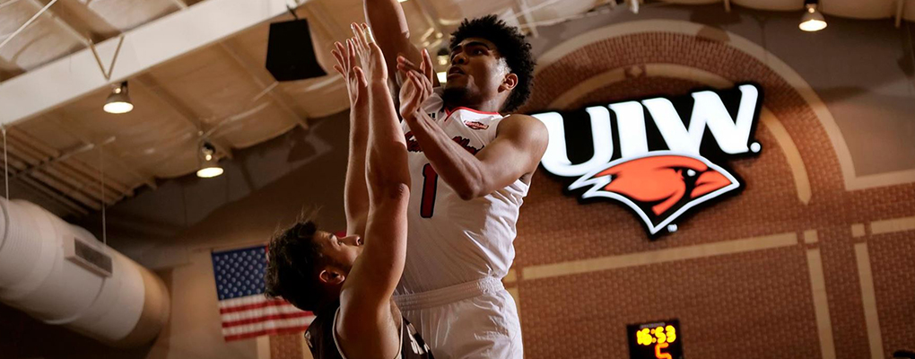 A UIW basketball player shoots his shot with the UIW logo illuminated behind him