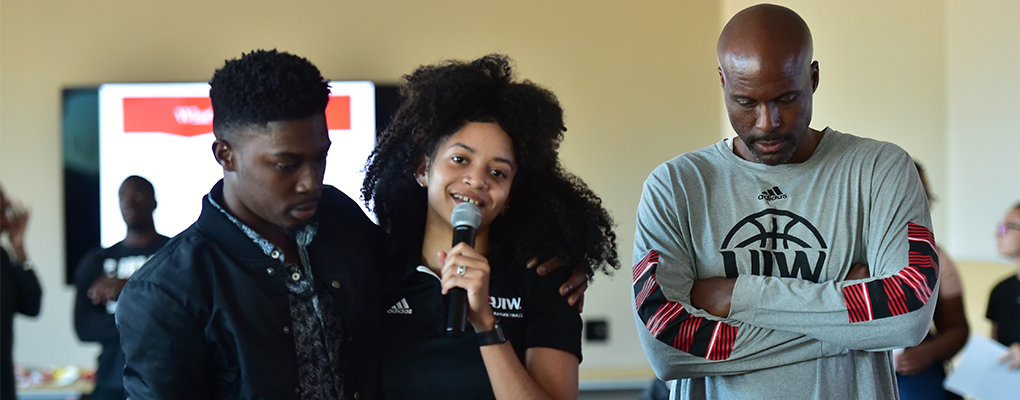 A student speaks into a microphone as she drapes her arm around her friend's shoulders