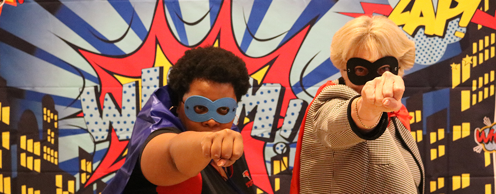 UIW employees stand in front of a super hero themed photo backdrop. They are wearing masks and capes, and extended their arms in front of them in a superhero pose. 