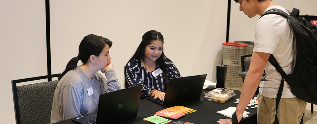 Two women sit behind an info table as a student stands on the other side