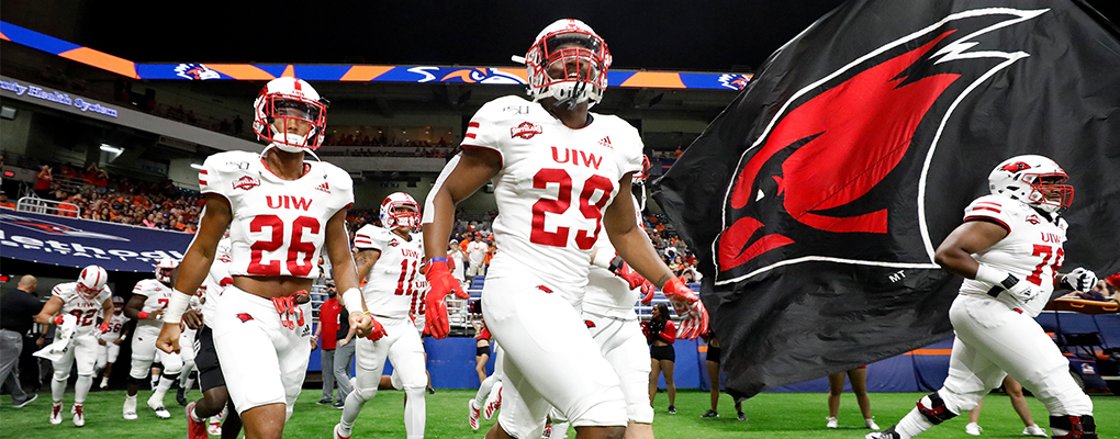 UIW football players run onto the football field with a giant red and black flag with an image of a Cardinal head