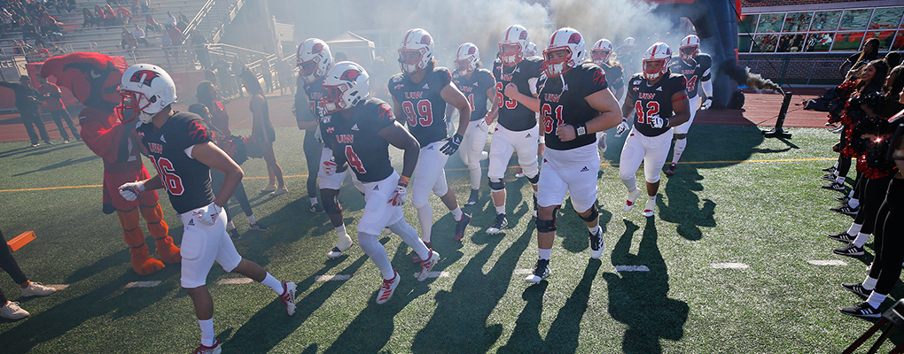 UIW Football players run onto the field through a cloud of smoke surrounded by cheerleaders