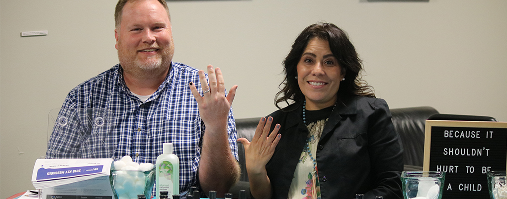 A man and a woman sit behind a table and hold up their hands to show a polished finger nail