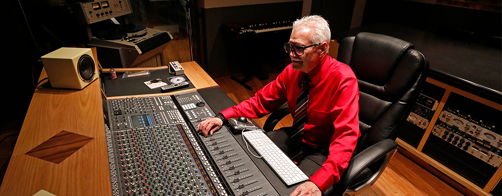 A man (Jim Waller) wearing a red, button down shirt sits behind a sound mixing board in a recording studio