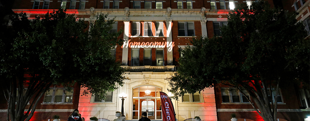 The words "UIW Homecoming" are illuminated on the outside wall of a red brick building