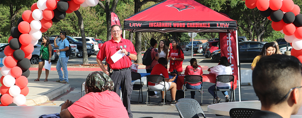 A man stands in front of a red and black balloon arch holding a microphone to address a crows