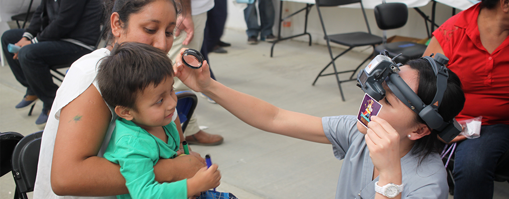 A UIW volunteer performs an eye exam on a little boy sitting on his mother's lap