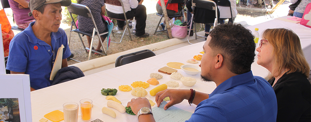 UIW volunteers sit with a man who listens to them speak. On the table in front of them are samples of healthy foods. 