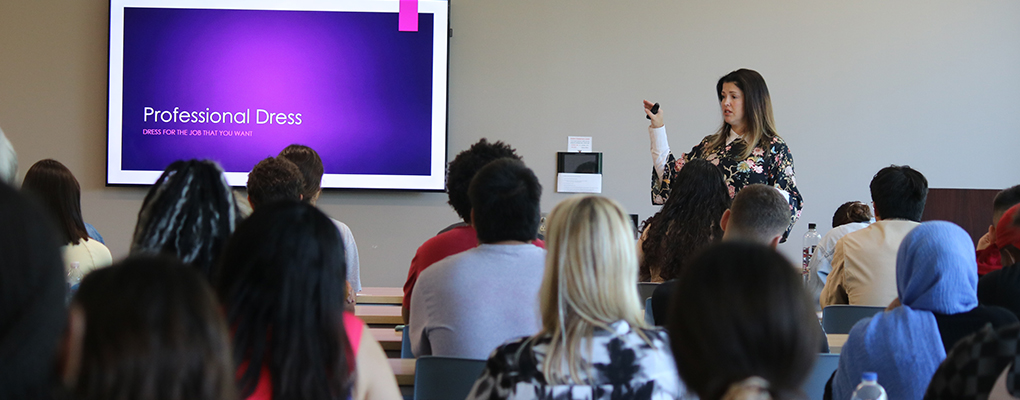 A woman stands at the front of a classroom and speaks as students sitting listen