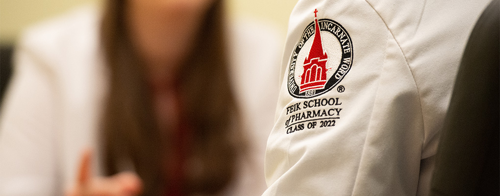 A white coat sleeve with a patch of the UIW Feik School of Pharmacy logo 