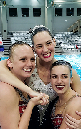Three synchronized swimming team members hug and pose for a photo in front of a swimming pool