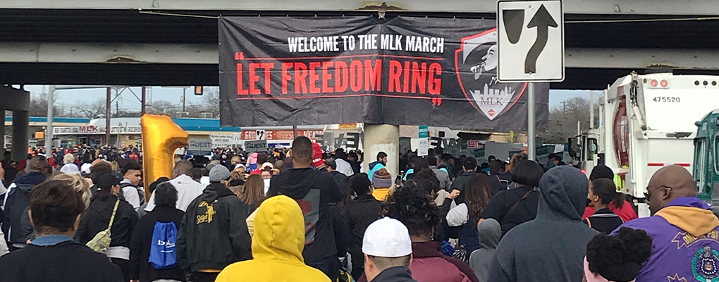 A crowd of people walk past a sign about the MLK March