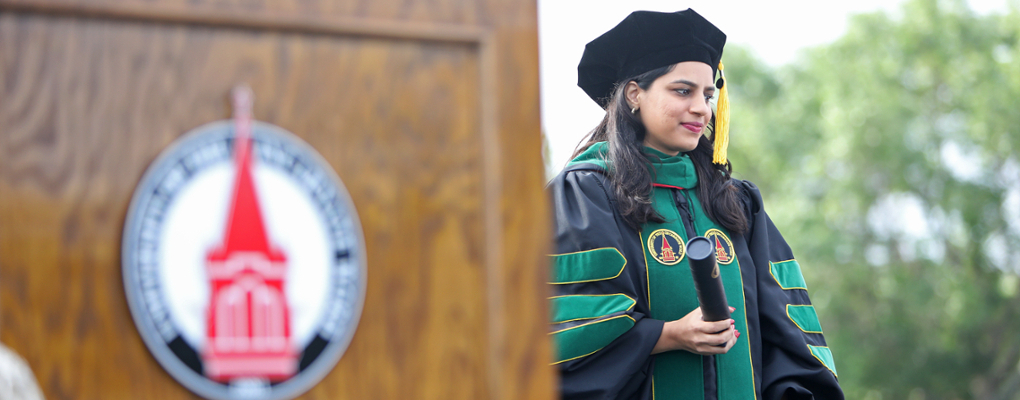 A doctoral student stands on stage in academic regalia