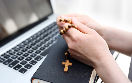 Hands in prayer over a Bible with a rosary in front of a laptop
